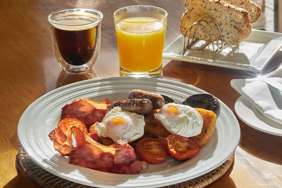 A breakfast plate with eggs, bacon, sausages, and tomatoes. There are two glasses of beverages and a slice of bread on the table.