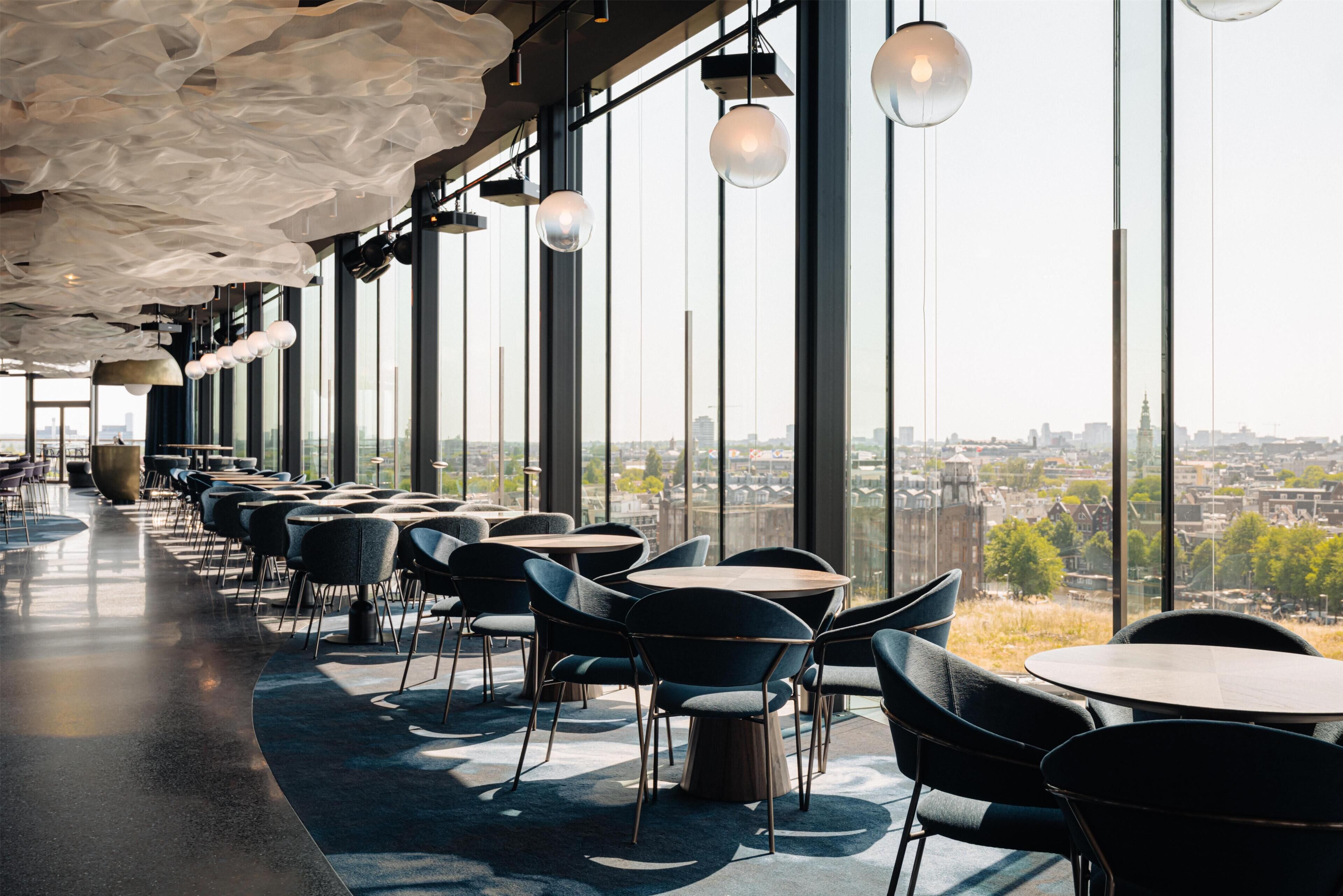 A modern interior with a long row of tables and chairs, large windows offering a city view, and unique ceiling design.