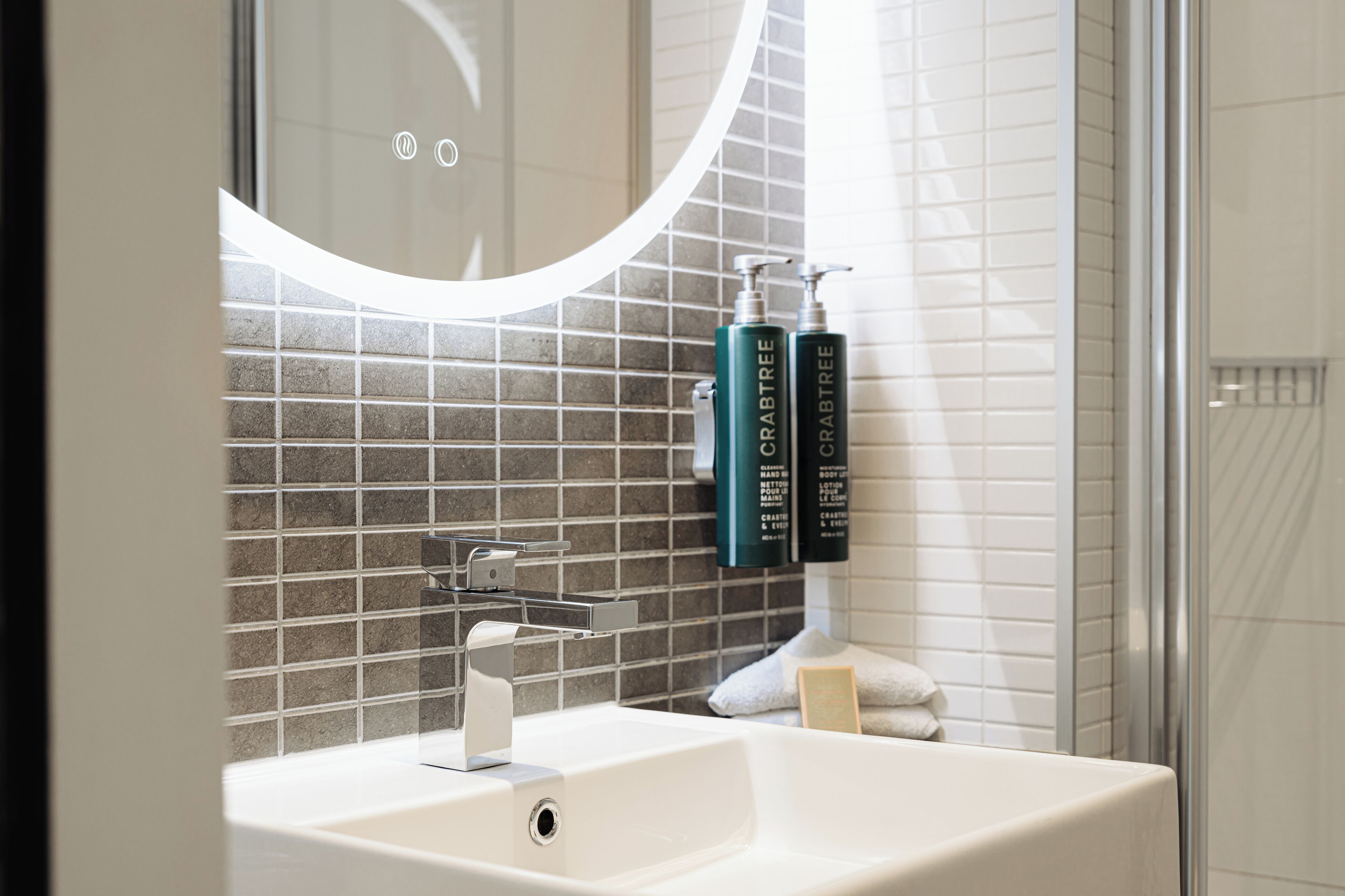 A bathroom with a white sink, a mirror with a circular light, and two bottles of Crabtree body wash on the wall.