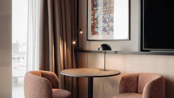 Een moderne kamer met een ronde tafel, twee gestreepten stoelen, een groot kunstwerk in een lijst en uitzicht op een stadsgezicht door een raam.