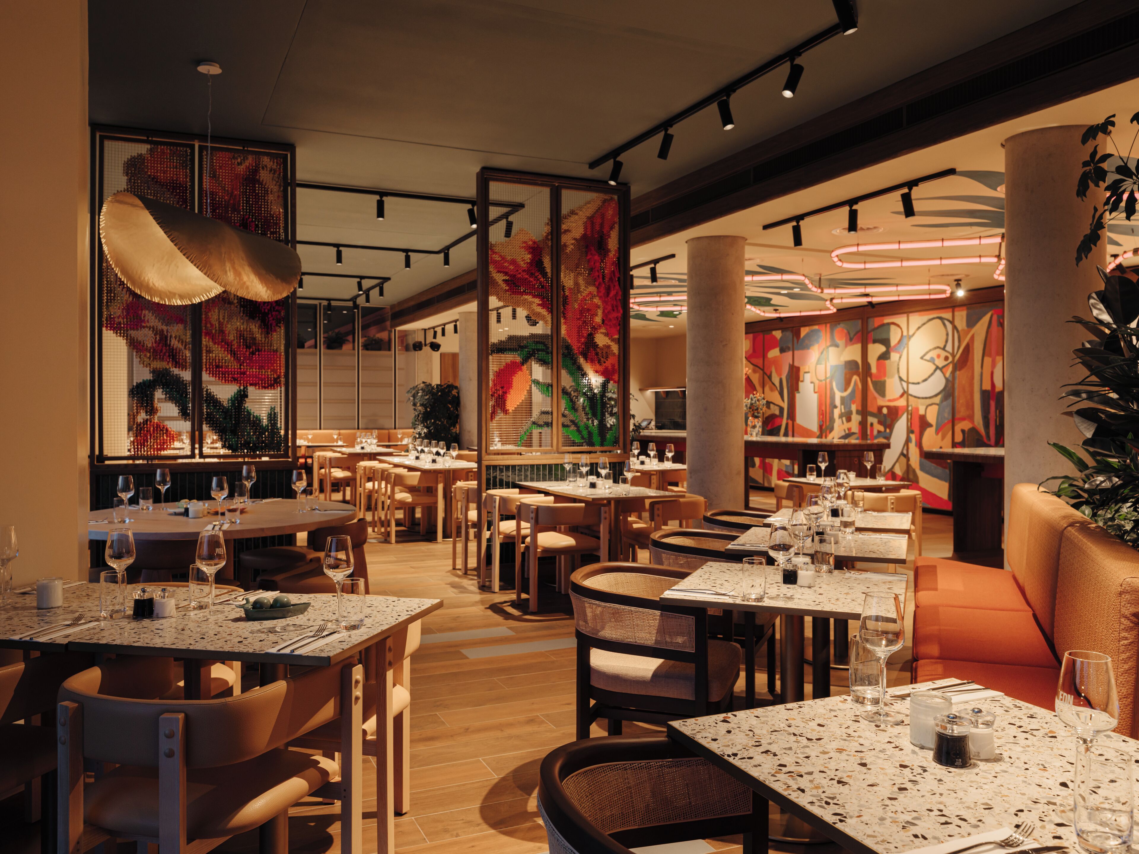 Un restaurante con varias mesas y sillas, una planta decorativa grande y un mural abstracto colorido en la pared.