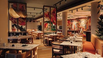 Un restaurante con varias mesas y sillas, una planta decorativa grande y un mural abstracto colorido en la pared.