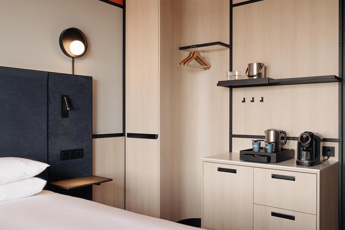 A hotel room with a bed, bedside table, a wall-mounted light, a wardrobe, a small shelf with a coffee maker, and a hanging rack.