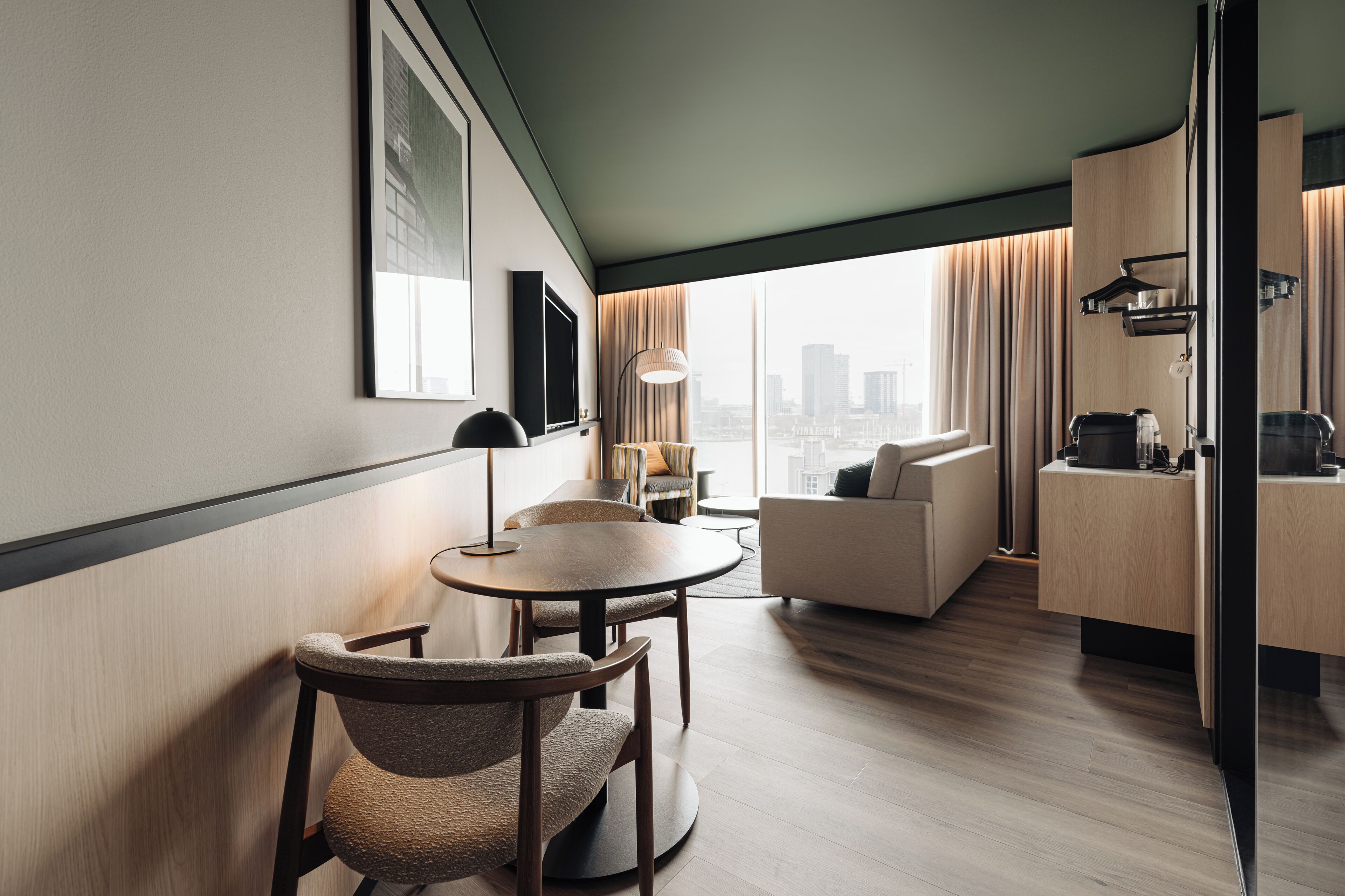 A modern hotel room with a dining area, a sofa, a kitchen counter, and a large window with a city view.