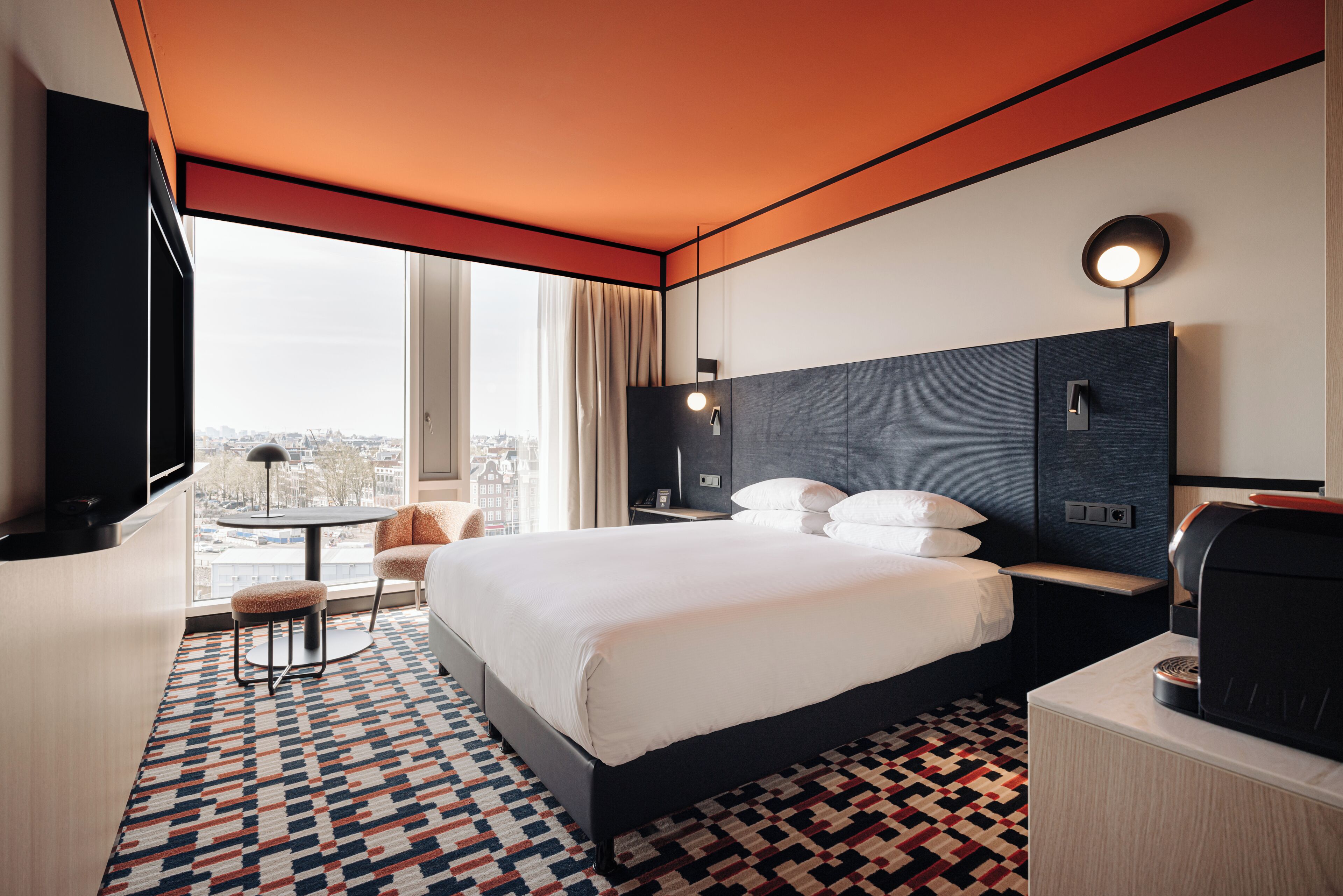 A hotel room with a large bed, a desk with a coffee machine, a chair, a round table, and a view of the city through the window.