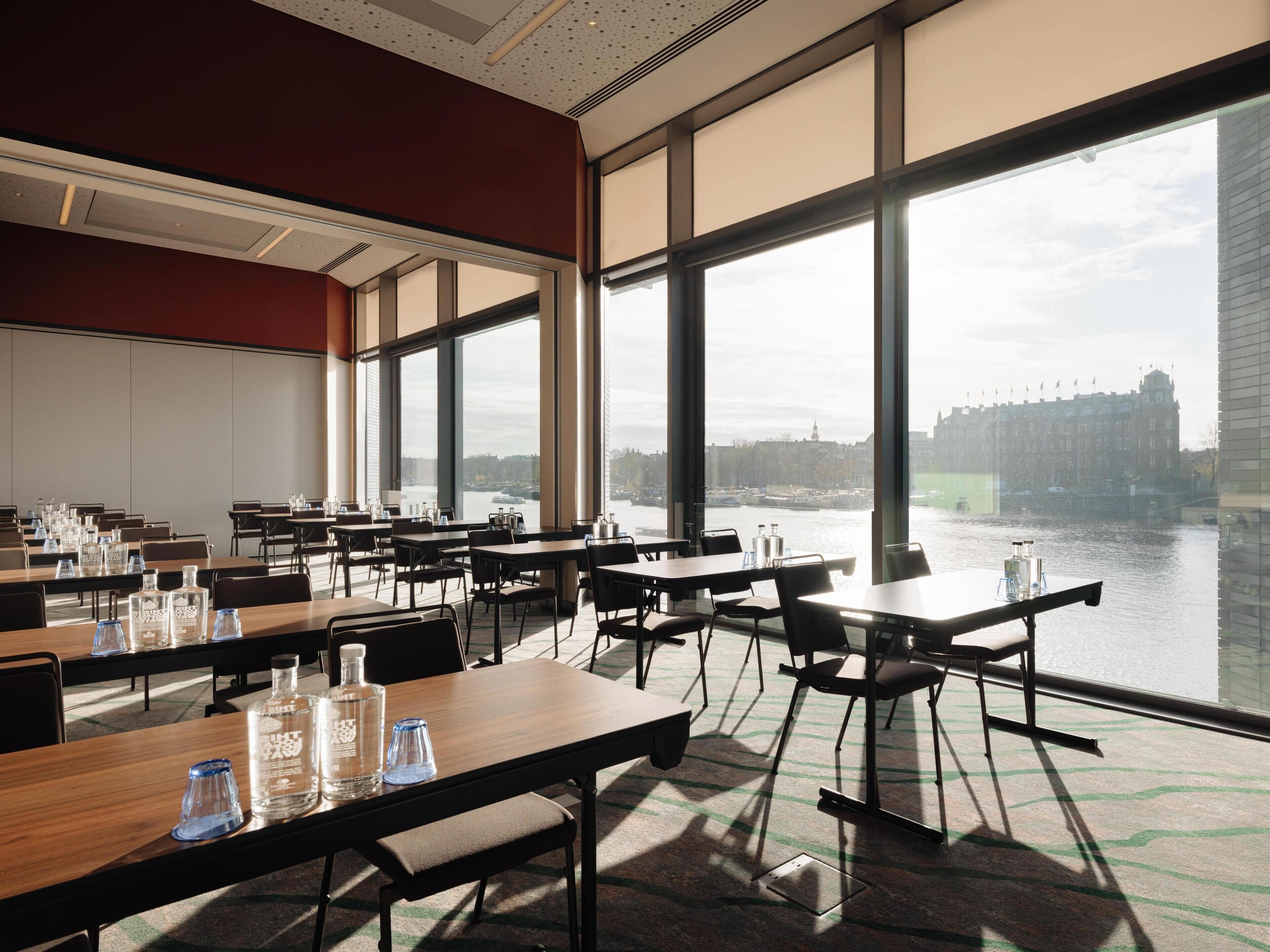 A conference room with tables and chairs, large windows with a city view, and a red accent wall.