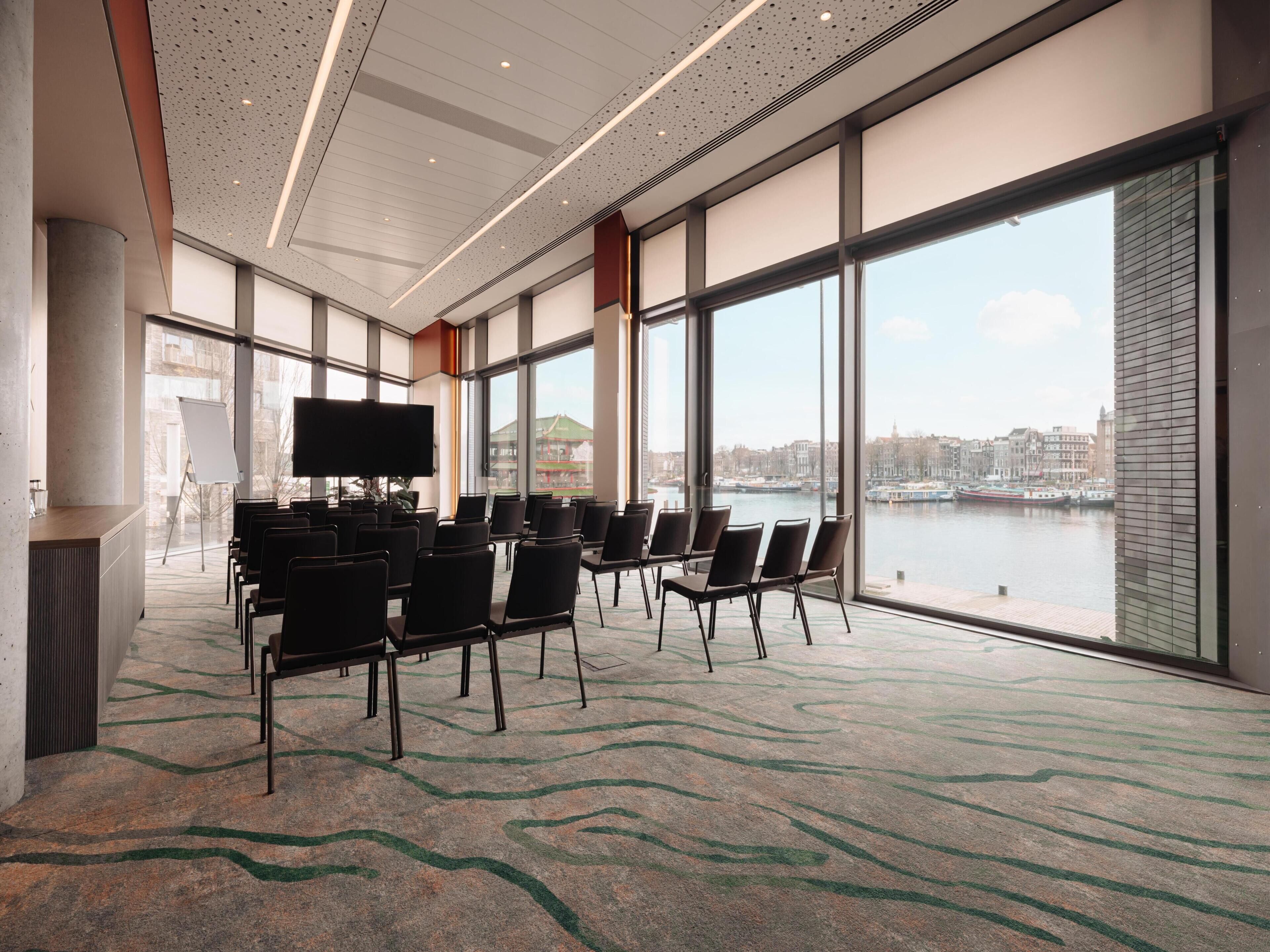 Une salle de conférence dotée d’un grand écran, de chaises disposées en rangées et offrant une vue sur une rivière et le paysage urbain grâce à de grandes fenêtres.