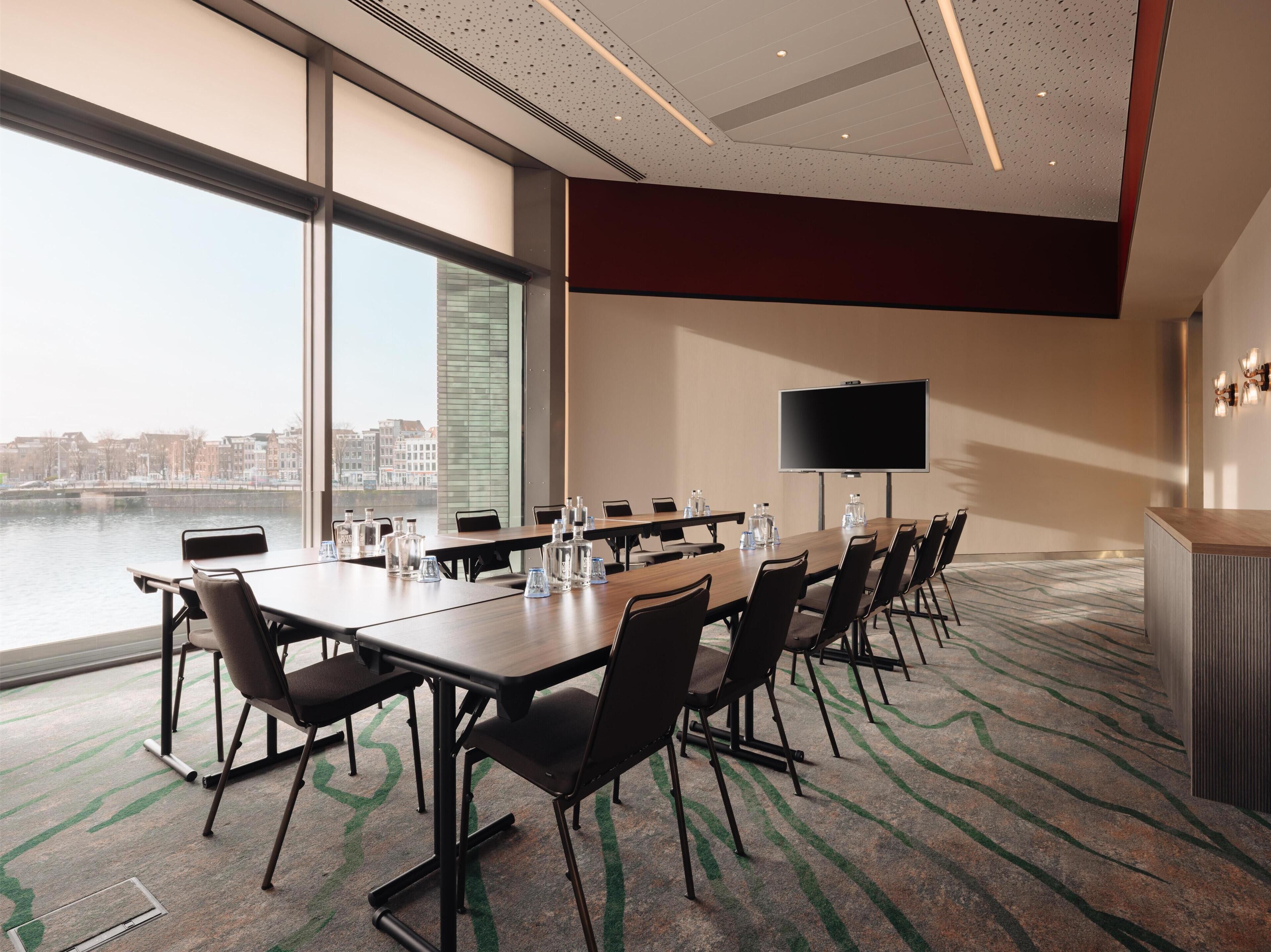 A conference room with a long table, chairs, a flat-screen TV, and a view of a cityscape through large windows.