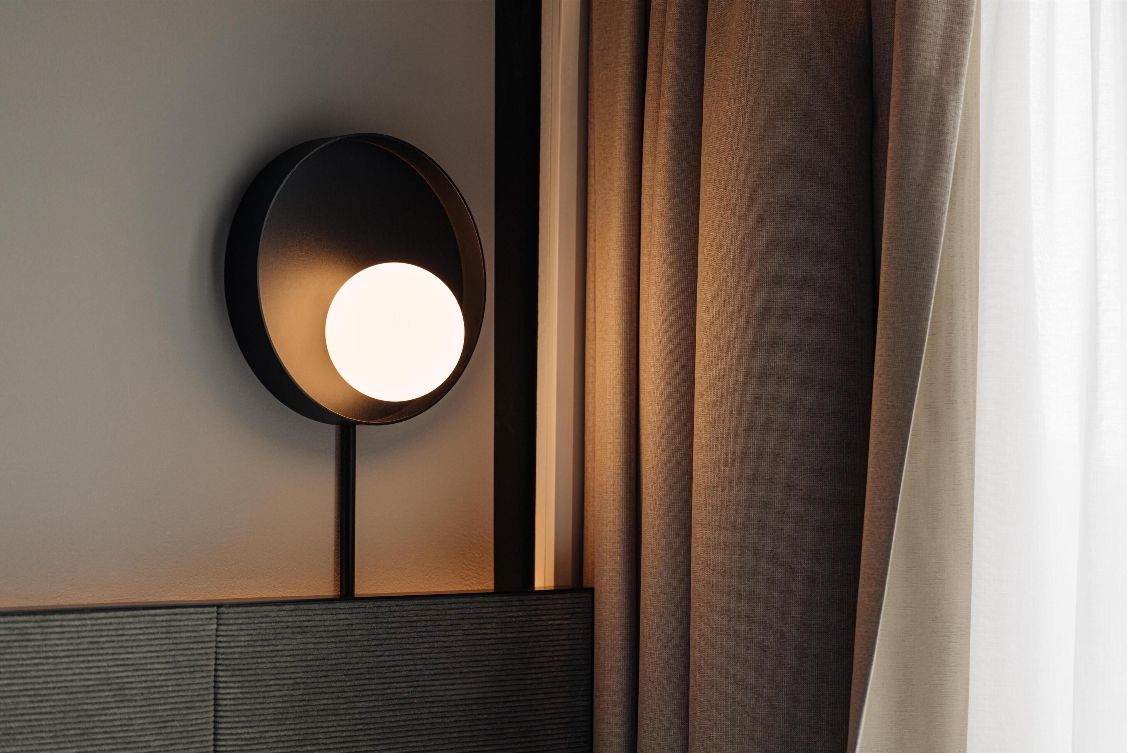 A wall-mounted light fixture with a circular shade, a vertical light pole, and a window with curtains.