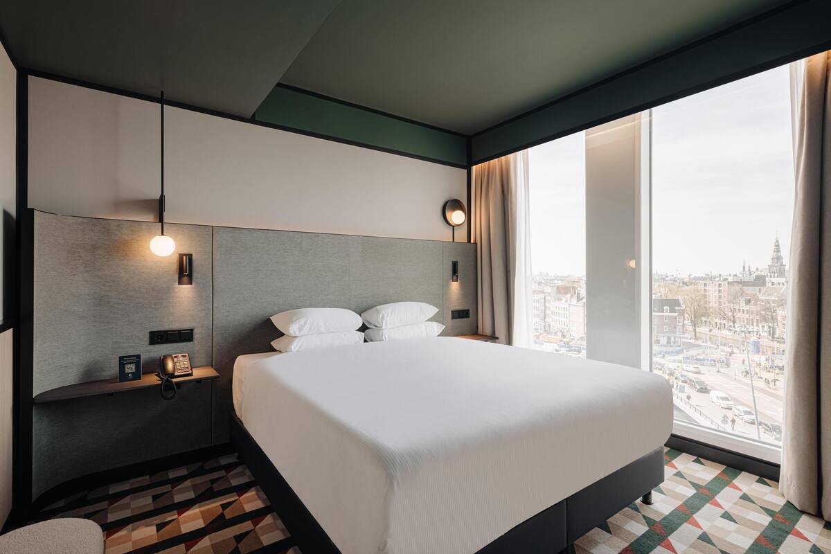 A modern hotel room with a large bed, a bedside table with a phone and a book, a wall-mounted light, and a city view through large windows.