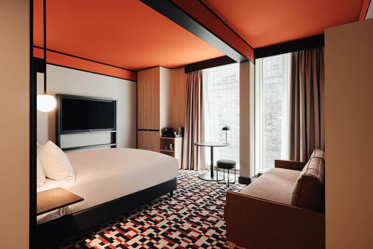 A hotel room with a bed, a TV, a desk, a chair, a small table, and a window with curtains.