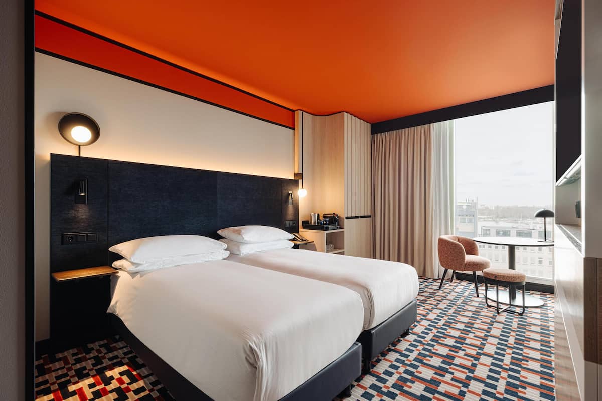 A hotel room with a large bed, bedside tables, a desk, a chair, and a view of the city.