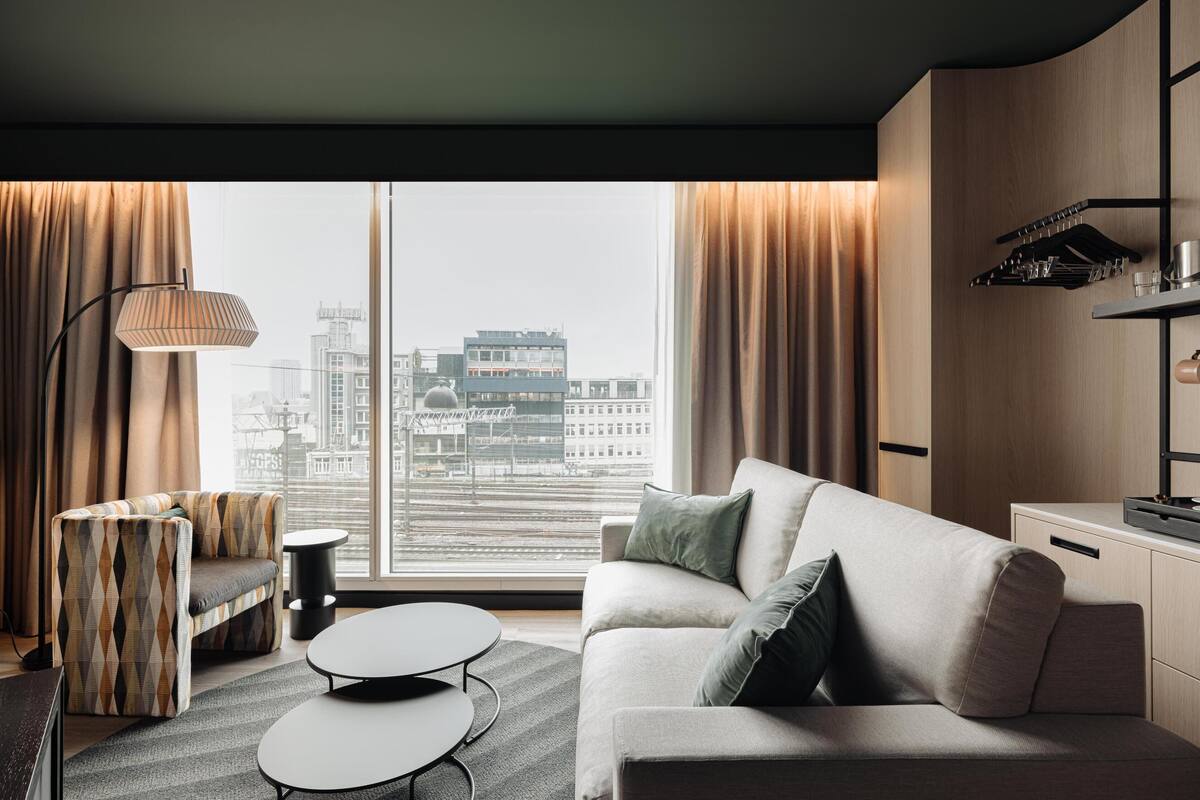 A modern hotel room with a sofa, armchair, and a dining table. A large window offers a view of the cityscape.