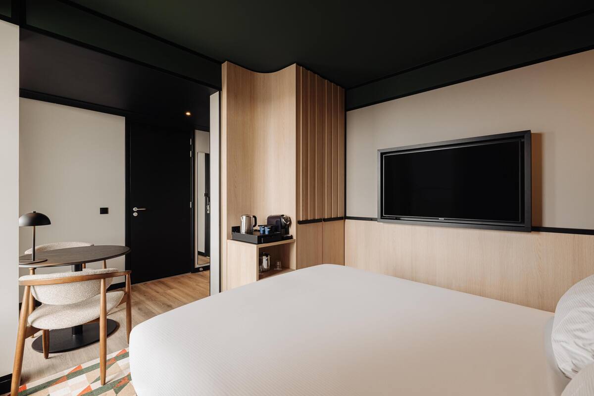 A hotel room with a bed, a TV, a desk with a chair, and a small kitchenette area.