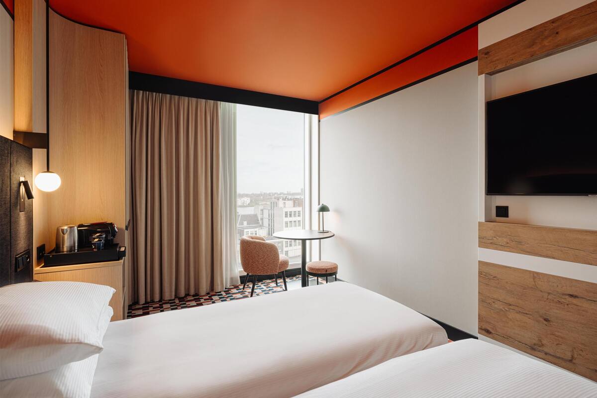 A hotel room with a bed, a desk with a coffee maker, a chair, a small table with a lamp, and a TV on the wall.