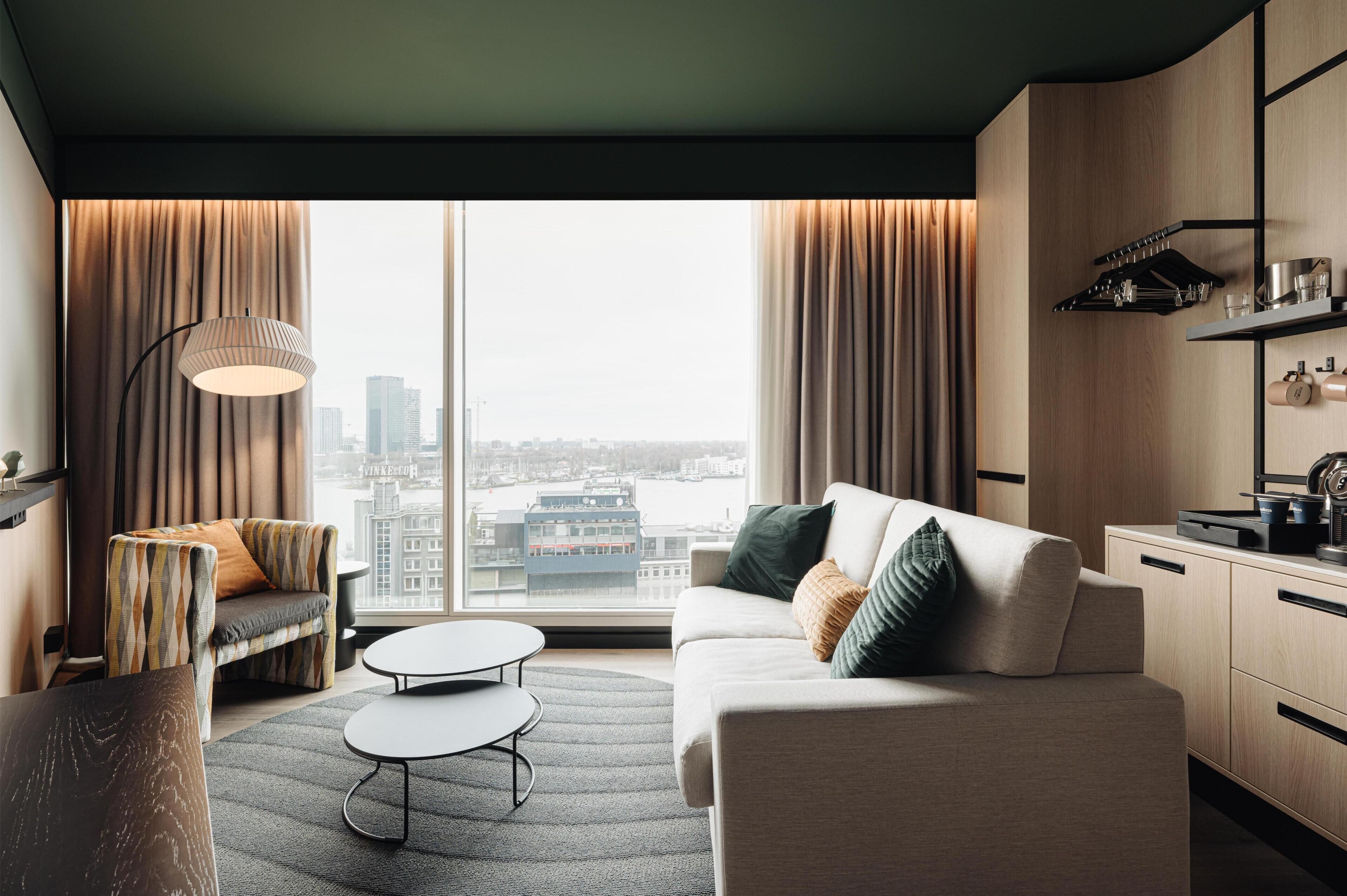 A modern hotel room with a sofa, armchair, dining table, and kitchen area.