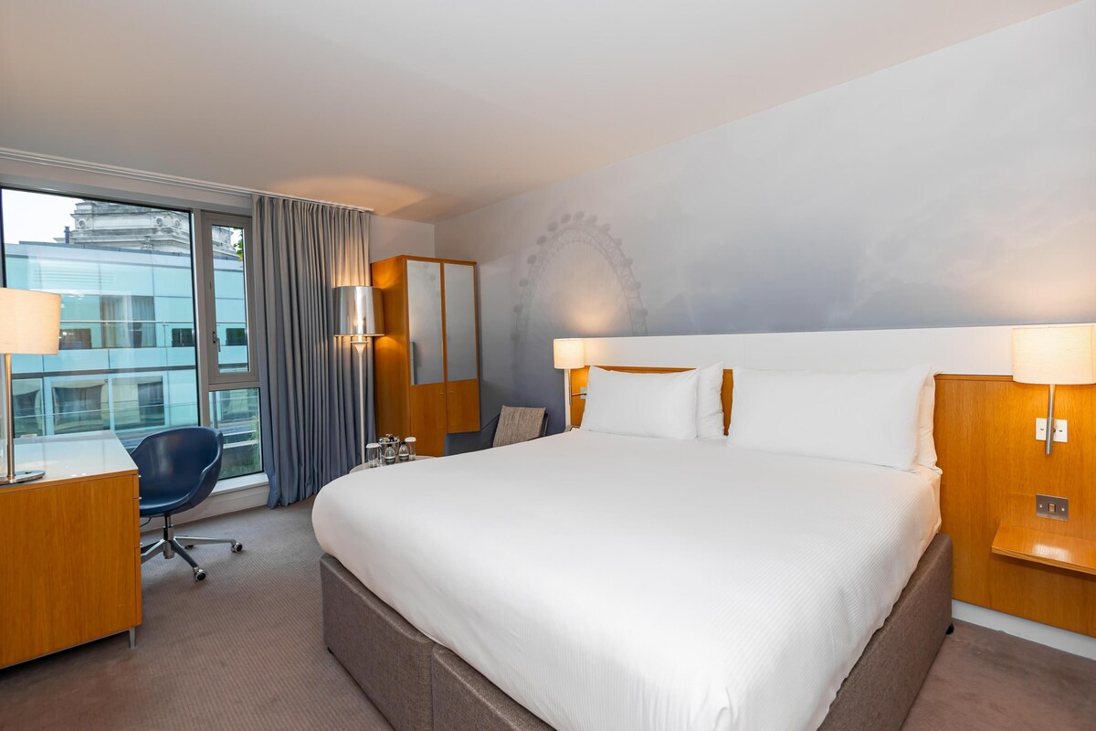A hotel room with a large bed, a desk with a chair, a lamp, and a view of a building through the window.