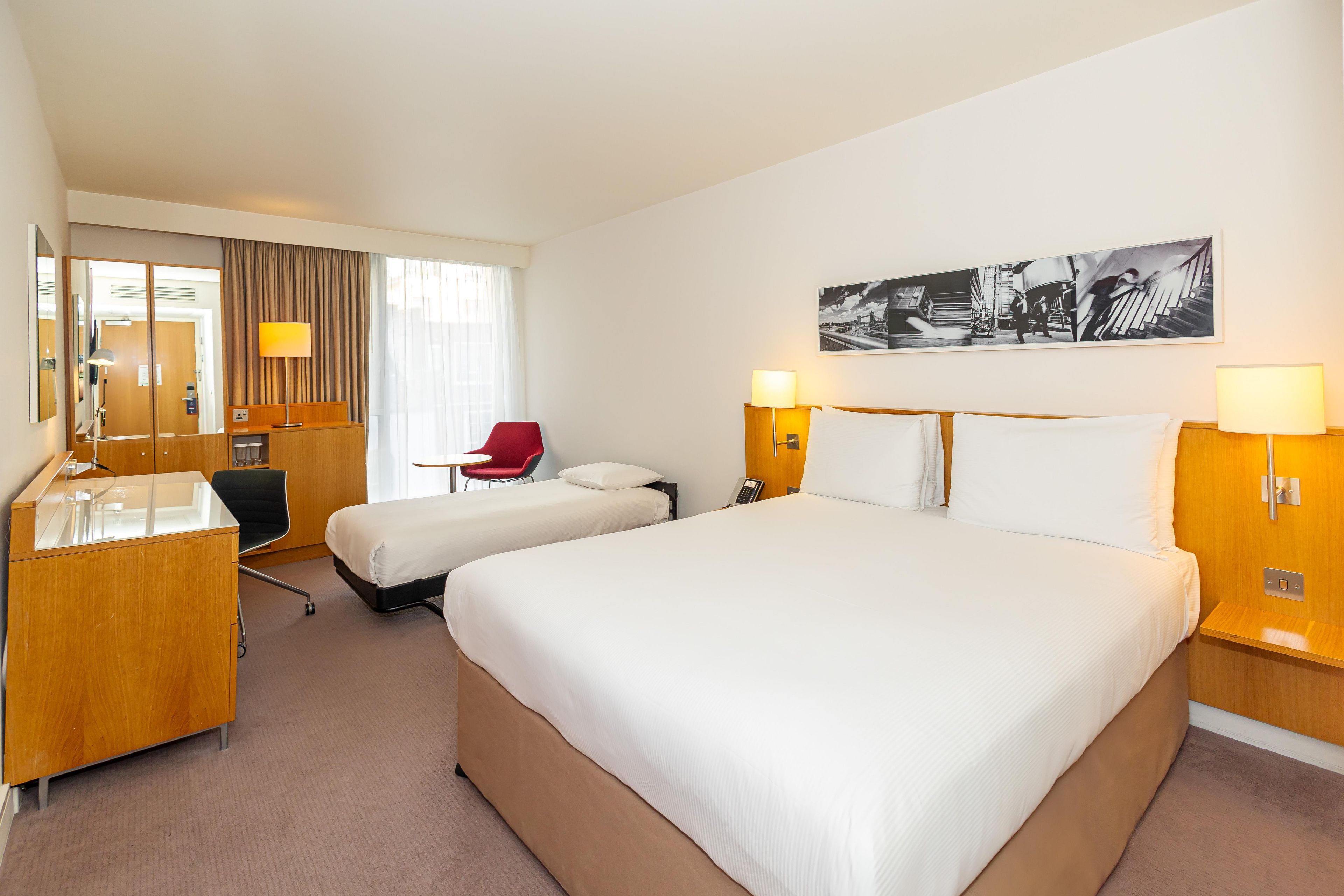 A hotel room with two beds, a desk, a chair, a telephone, and a painting on the wall.