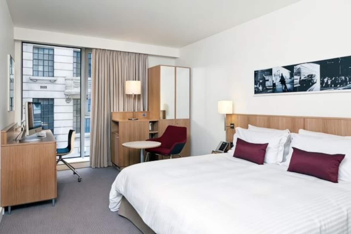 A hotel room with a bed, desk, chair, and a view of the city.