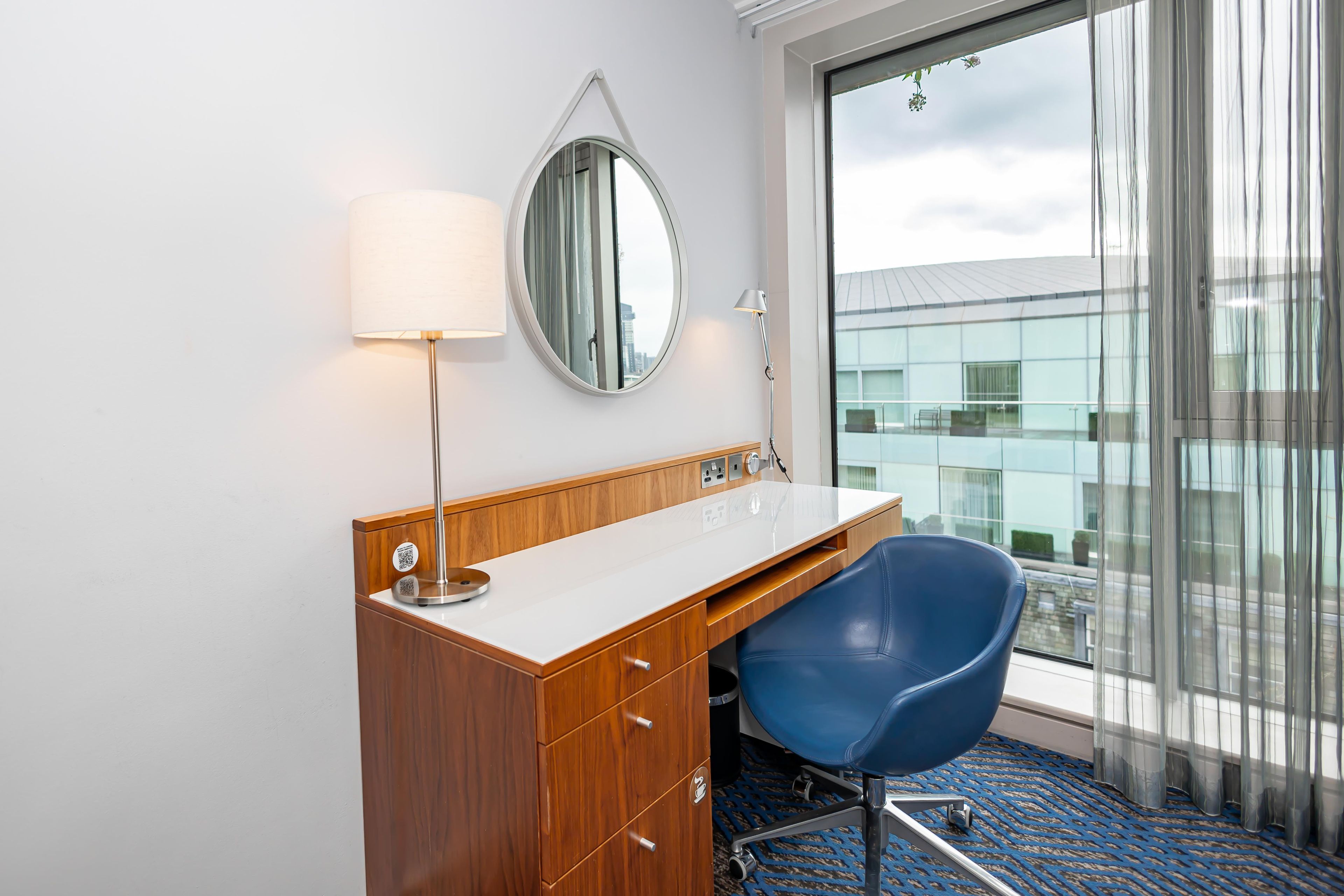 Ein Hotelzimmer mit einem Holztisch, einem blauen Sessel, einem Spiegel, einer Lampe und einem großen Fenster mit Blick auf Gebäude.