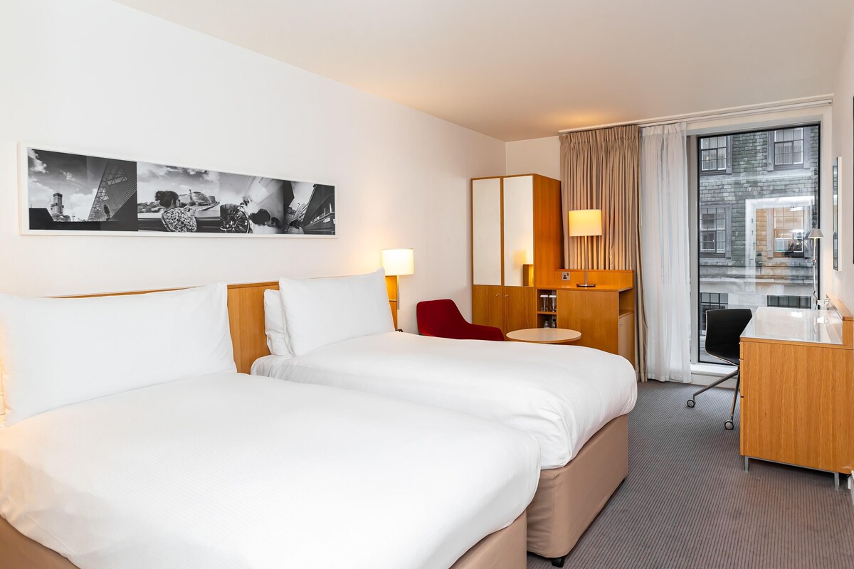 A hotel room with a large bed, a desk, a chair, a lamp, and a view of the city.