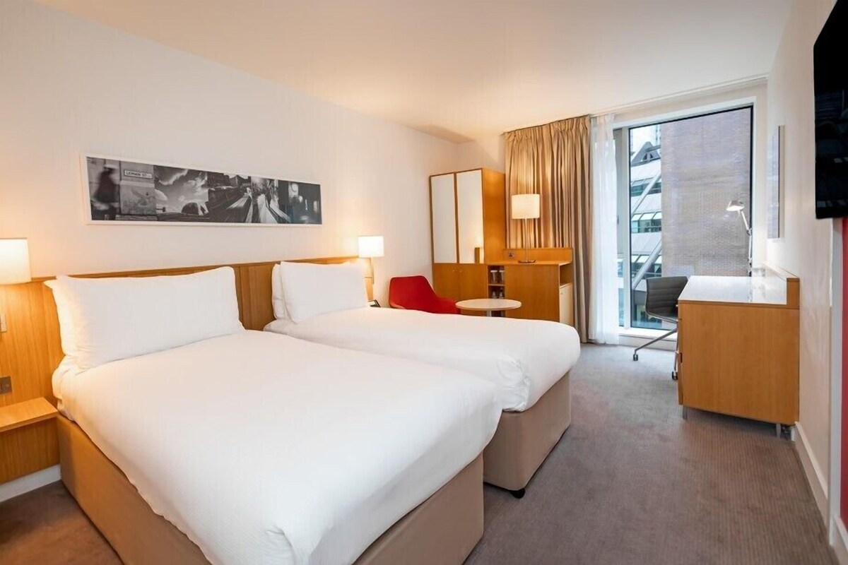 A hotel room with two beds, a desk, a chair, a TV, and a large window.