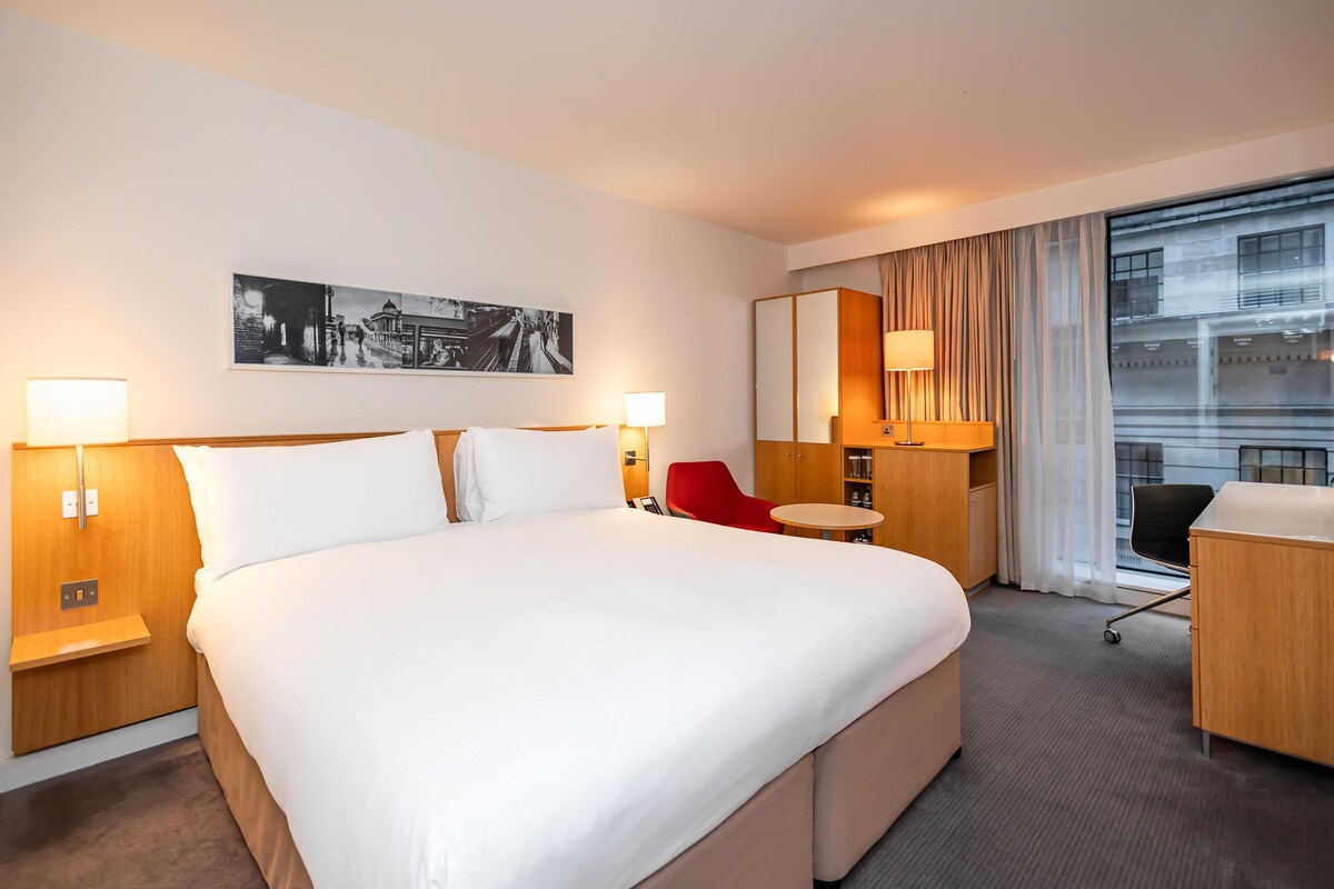 A hotel room with a large bed, bedside tables, a desk, a chair, a small round table, a red chair, a large window with curtains, and a wall-mounted picture.