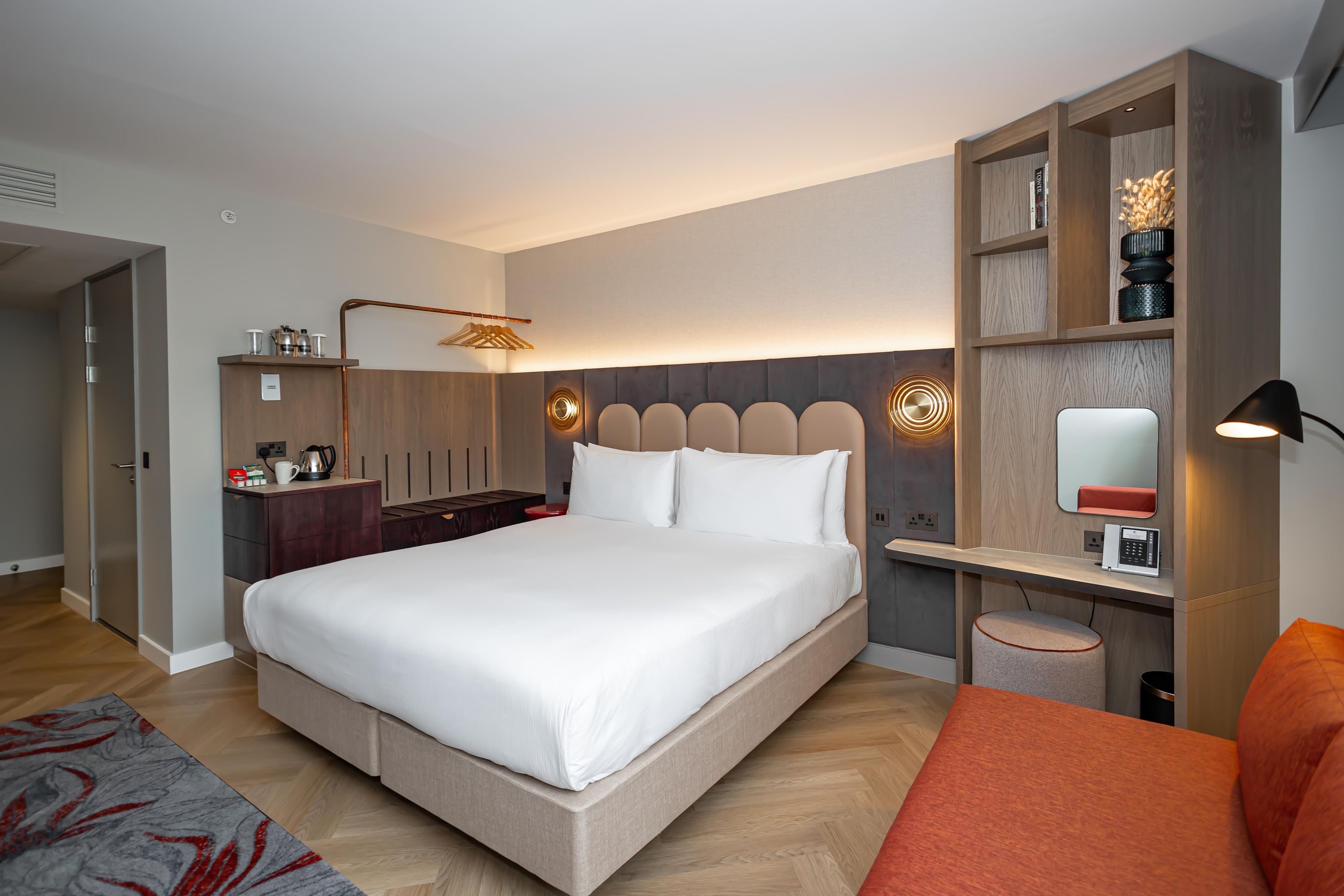 A modern hotel room with a large bed, a desk with a lamp, a small round table, and a bookshelf.
