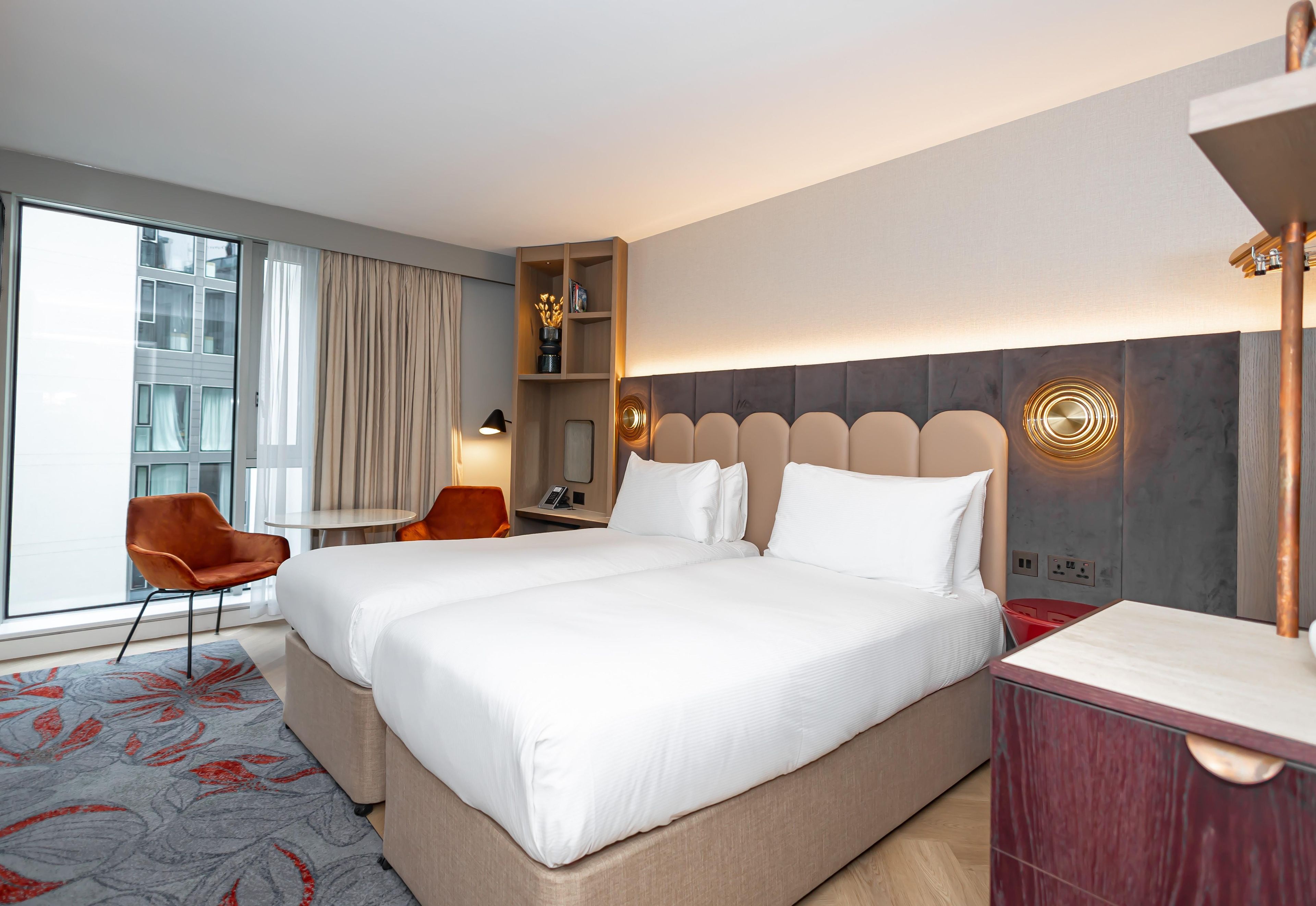 A hotel room with a large bed, two chairs, a desk, and a view of a building through the window.