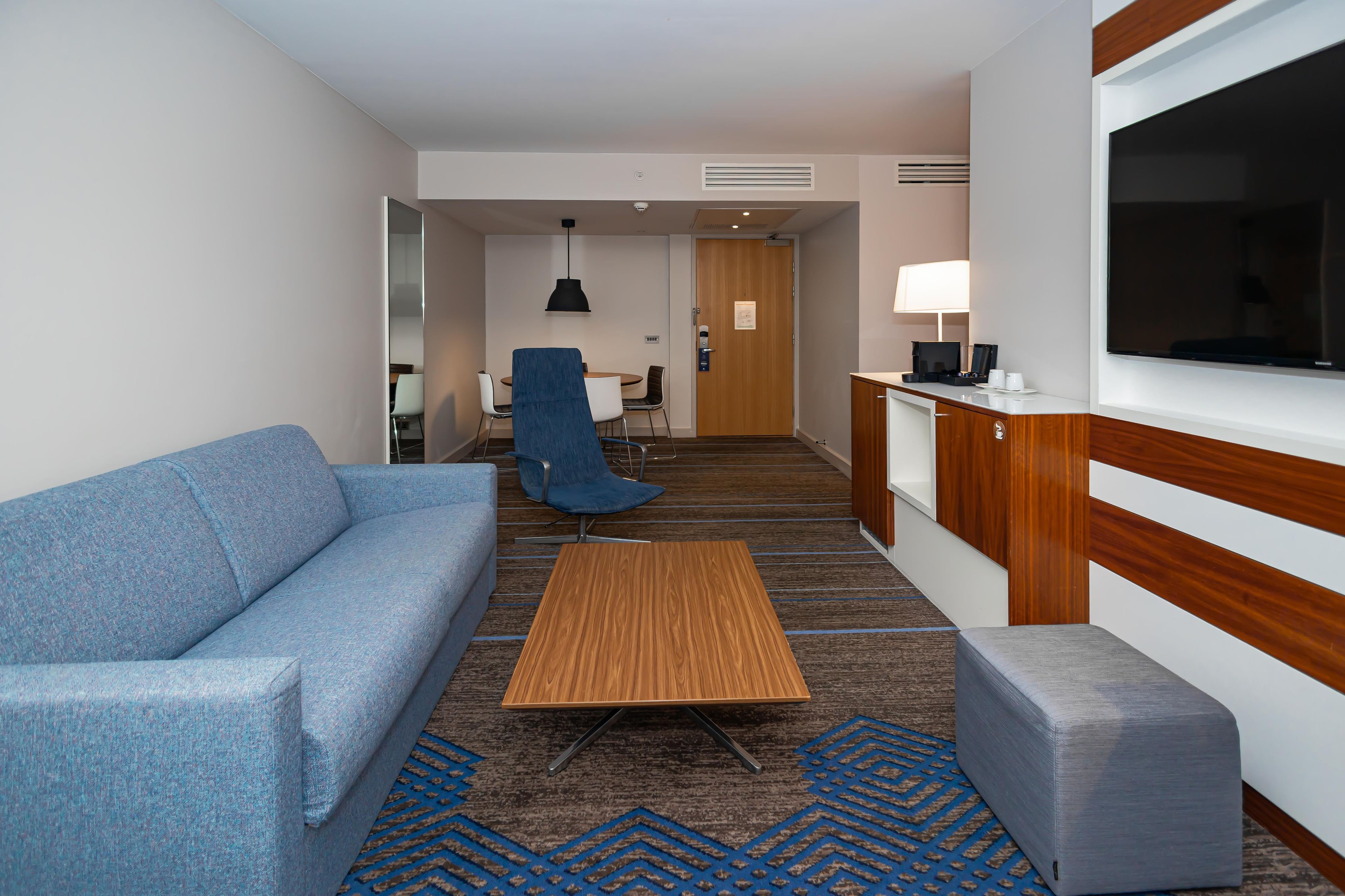 A hotel room with a sofa, a small table, a chair, a TV, a desk with a lamp, and a kitchenette area.