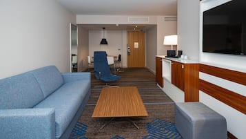 A hotel room with a sofa, a small table, a chair, a TV, a desk with a lamp, and a kitchenette area.