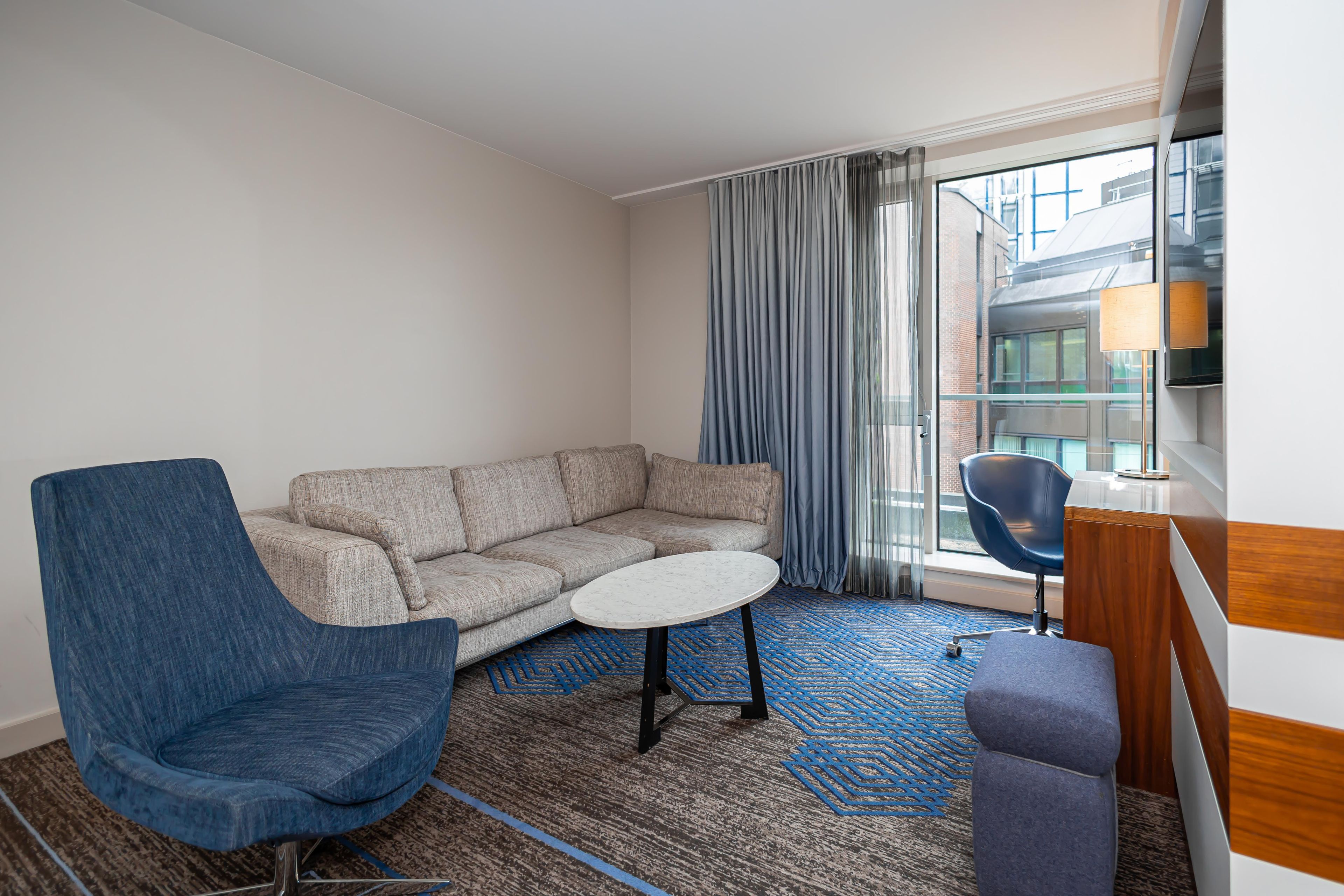 Ein modernes Hotelzimmer mit einer Eckcouch, einem runden Tisch, einem blauen Sessel und einem Schreibtisch mit Lampe.