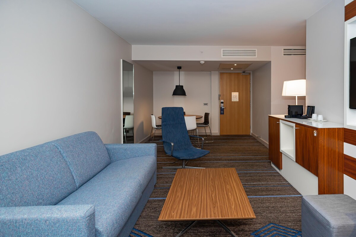 A hotel room with a blue sofa, a wooden coffee table, a blue armchair, a small desk with a chair, a lamp, and a TV on a wooden cabinet.