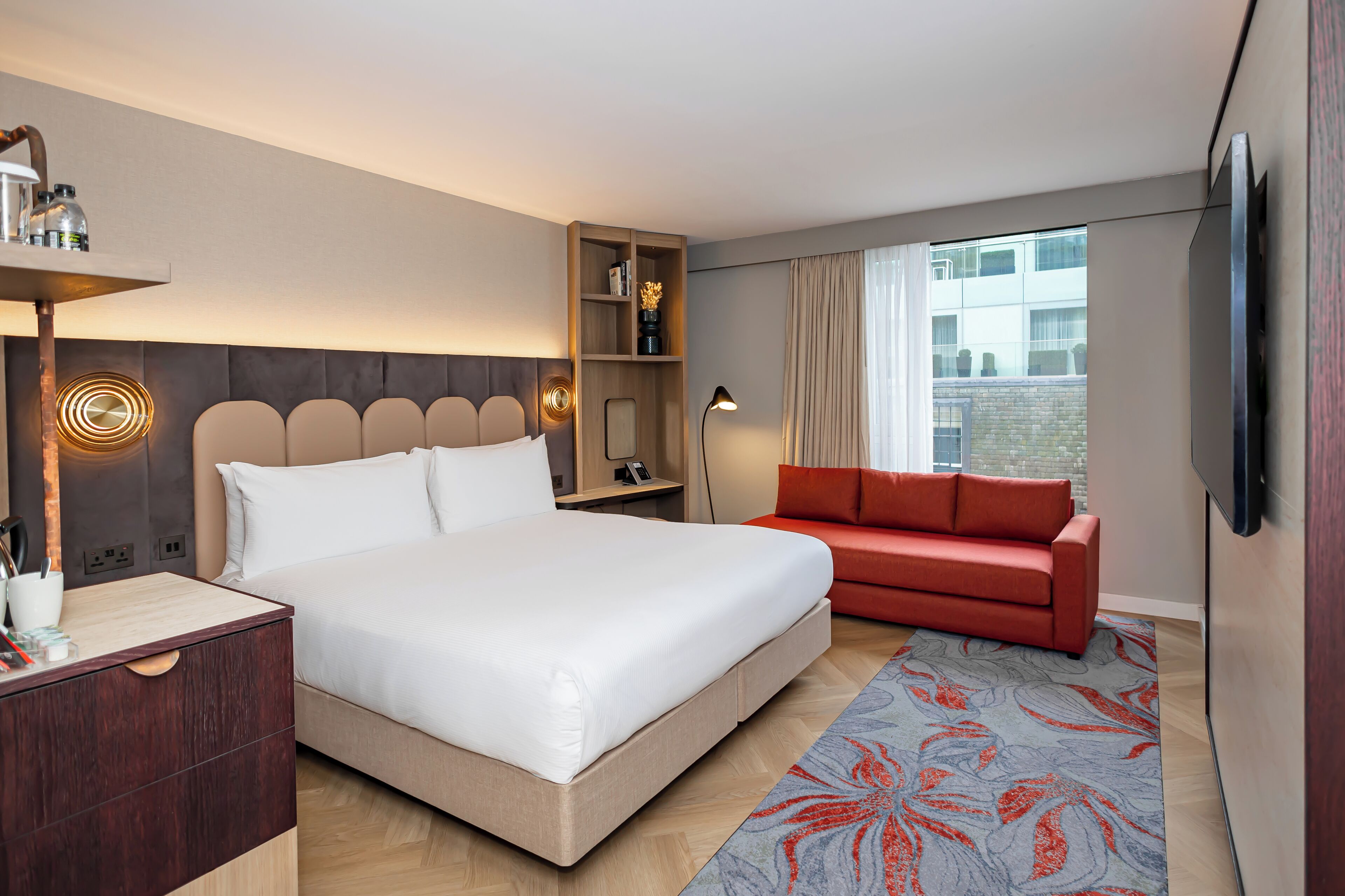A modern hotel room with a large bed, a red sofa, a TV mounted on the wall, and a wooden nightstand with a lamp.