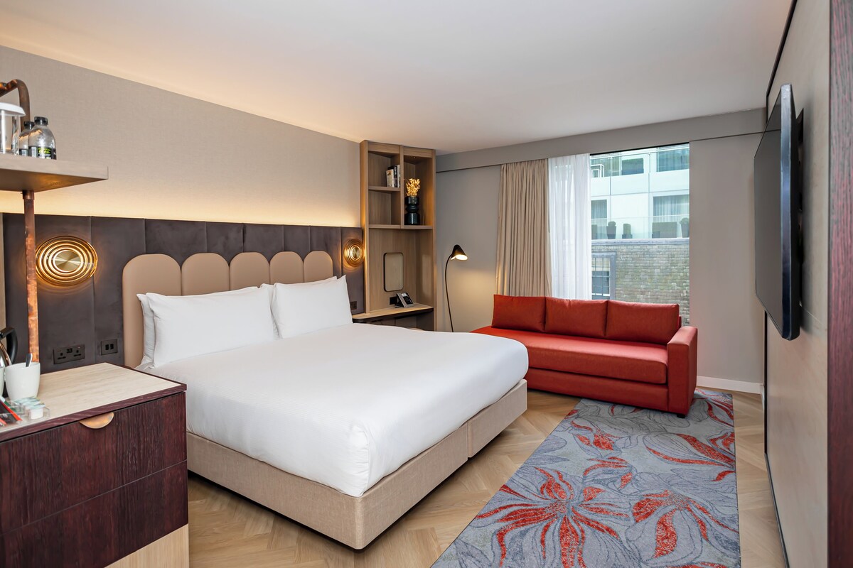A modern hotel room with a large bed, a red sofa, a TV mounted on the wall, and a wooden nightstand with a lamp.