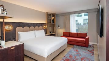 Ett modernt hotellrum med en stor säng, en röd soffa, en tv som är monterad på väggen och ett träbord vid sängen med en lampa.