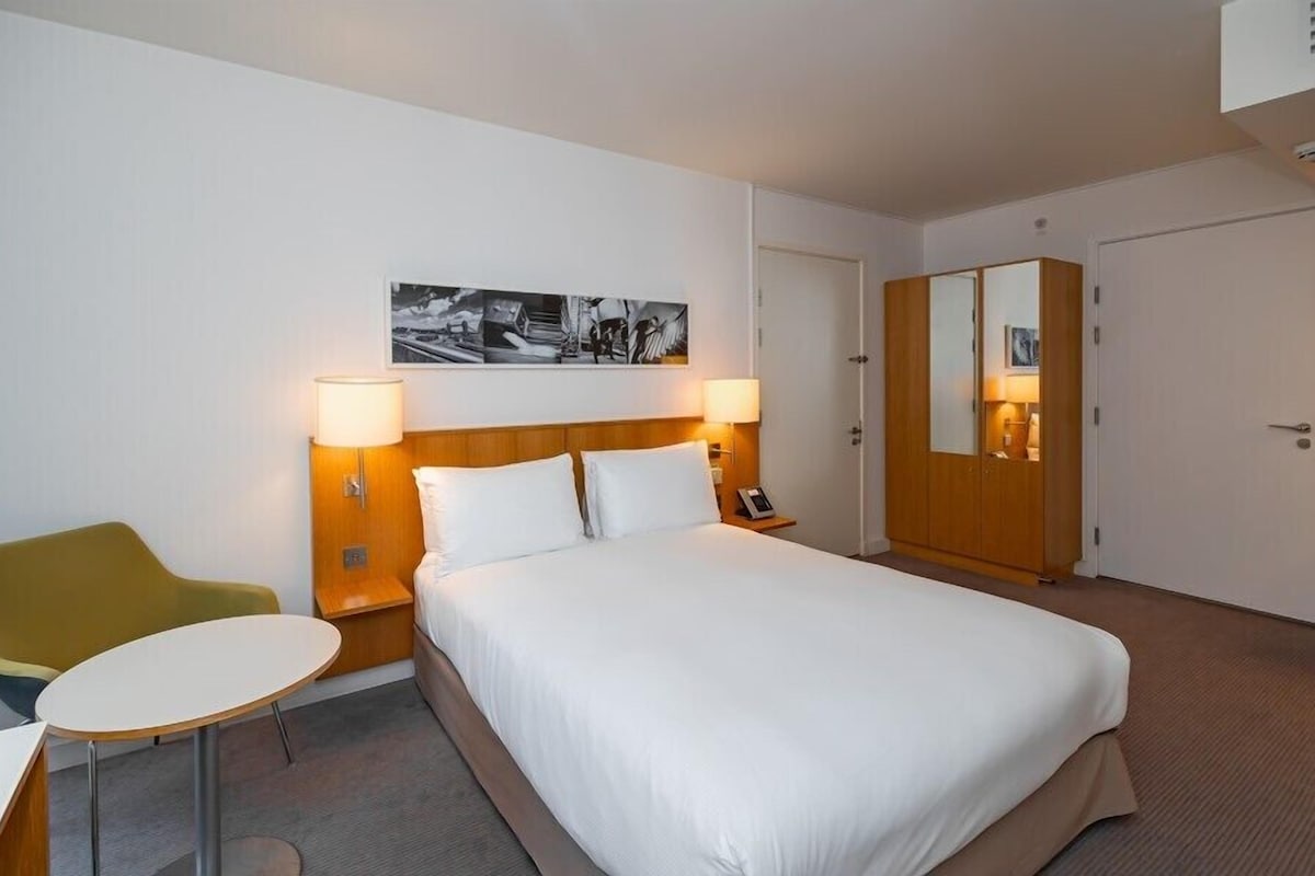 A hotel room with a bed, bedside tables, a chair, a small table, a wardrobe, and a wall-mounted artwork.