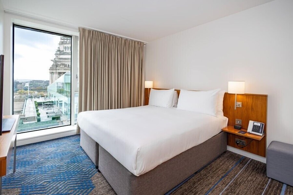 A hotel room with a large bed, a desk, a chair, a nightstand with a lamp, and a view of a building through a window.