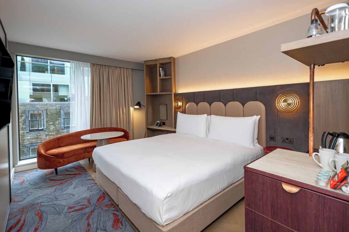 A hotel room with a large bed, a wooden nightstand, a desk, a chair, a bookshelf, and a window with a view of a building.