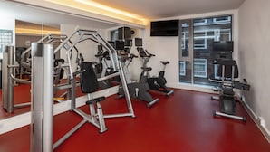A gym with exercise equipment, a large mirror, and a window with a view of buildings. - DoubleTree by Hilton Hotel London -Tower of London (London)