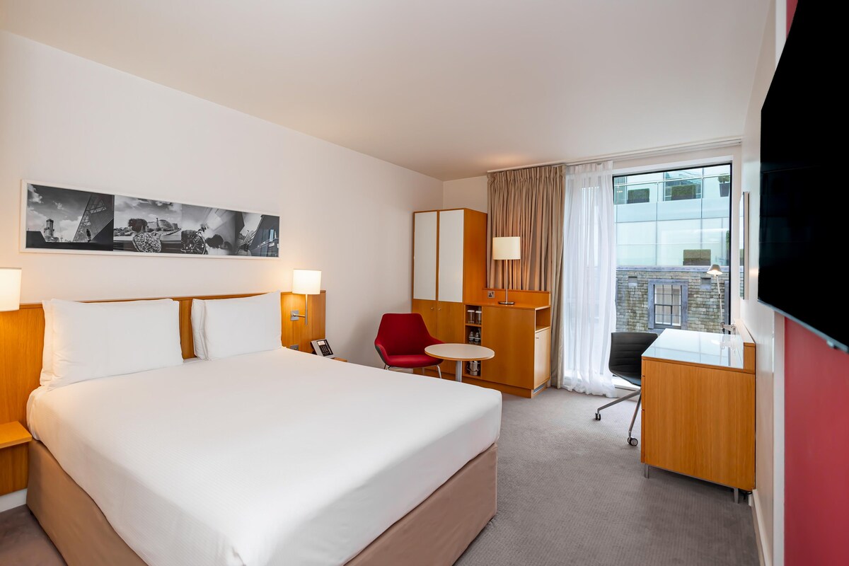 A hotel room with a large bed, a desk, a chair, a small table, a telephone, and a TV.