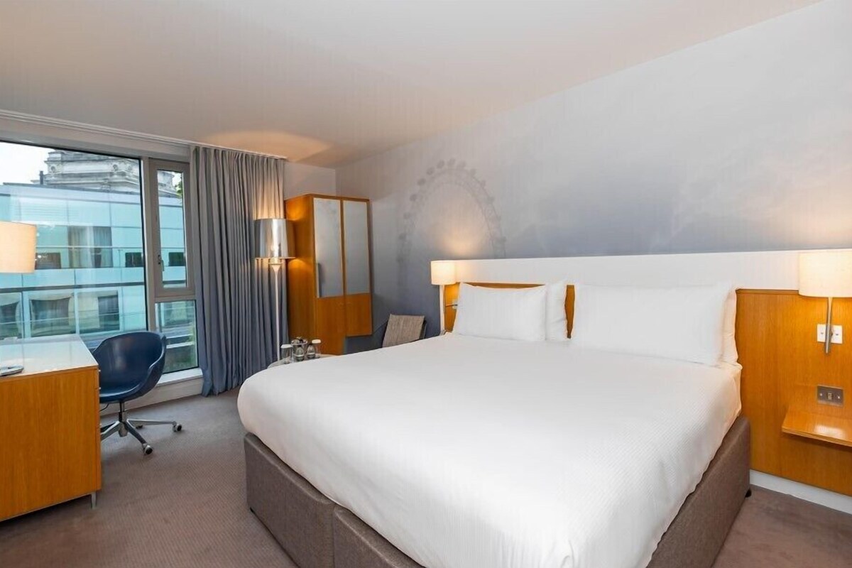 A hotel room with a large bed, a desk, a chair, and a view of the city through the window.