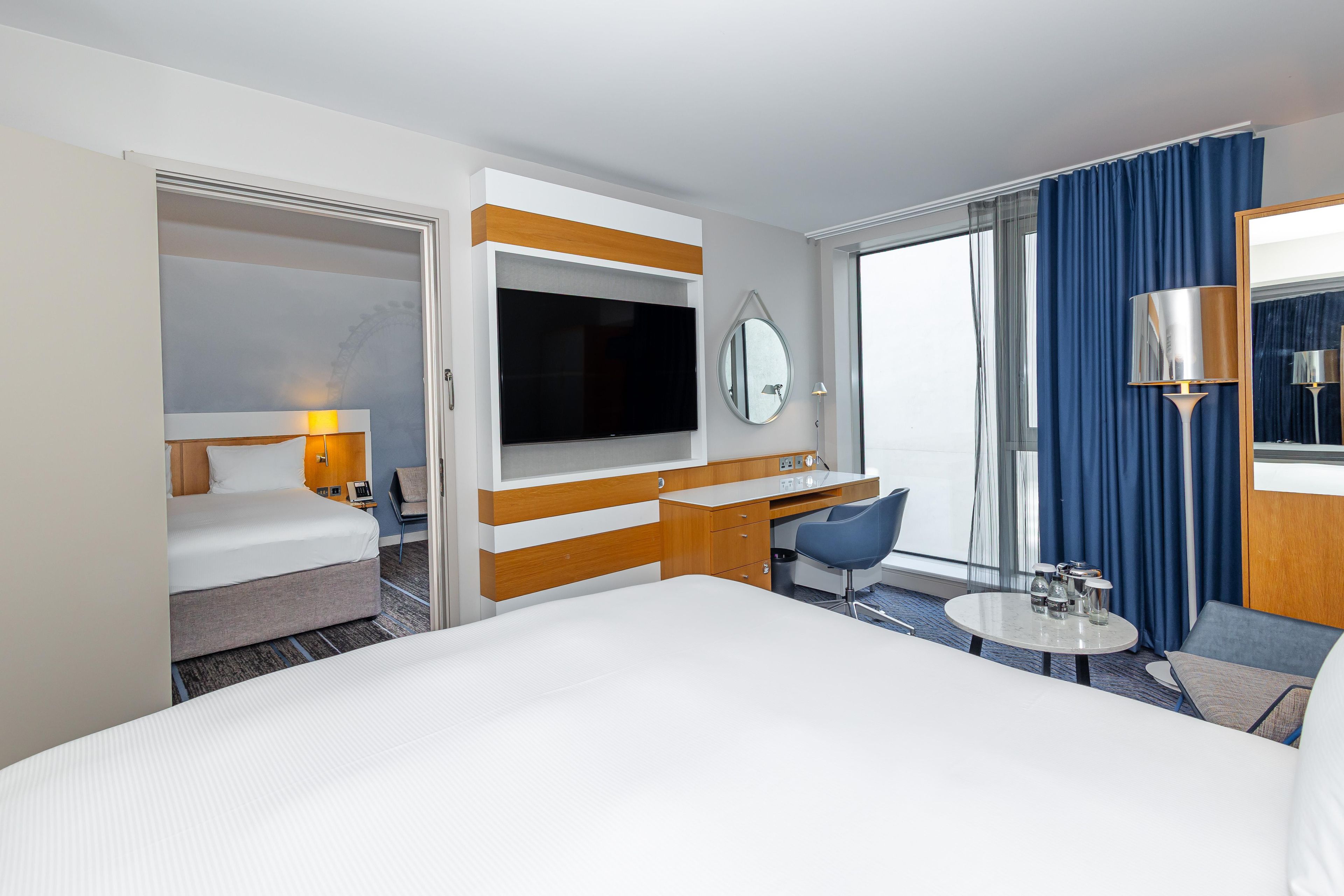 Habitación de hotel con una cama grande, un escritorio con silla, televisor, espejo y ventana con cortinas.