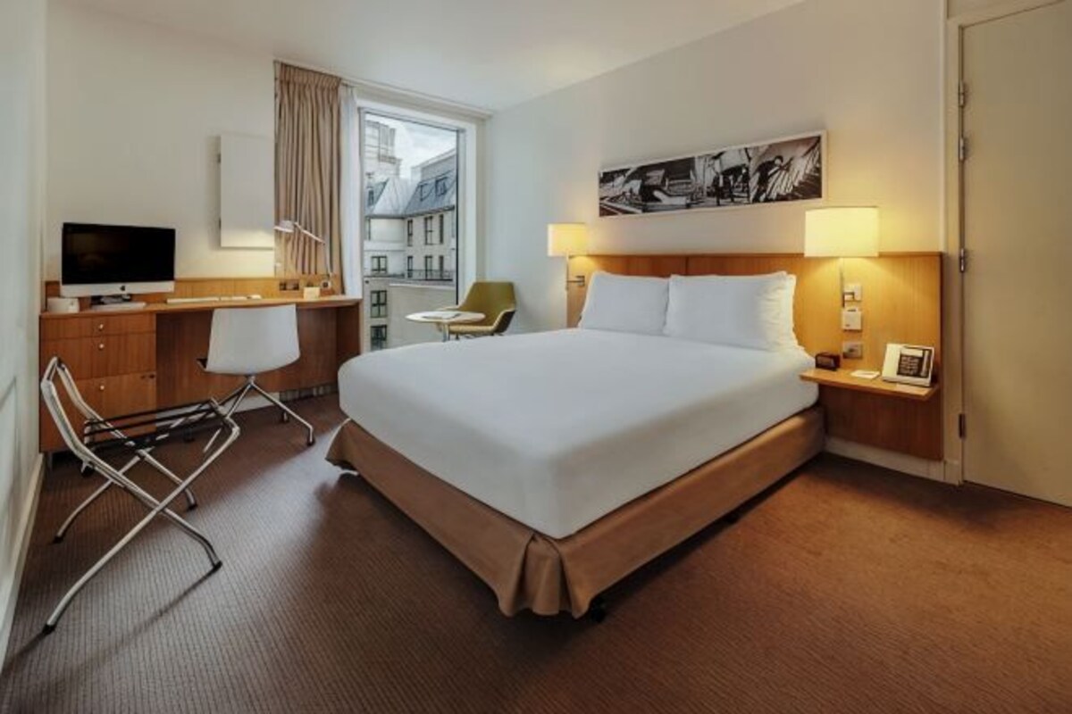A hotel room with a bed, a desk with a chair, a television, a window with curtains, and a city view.
