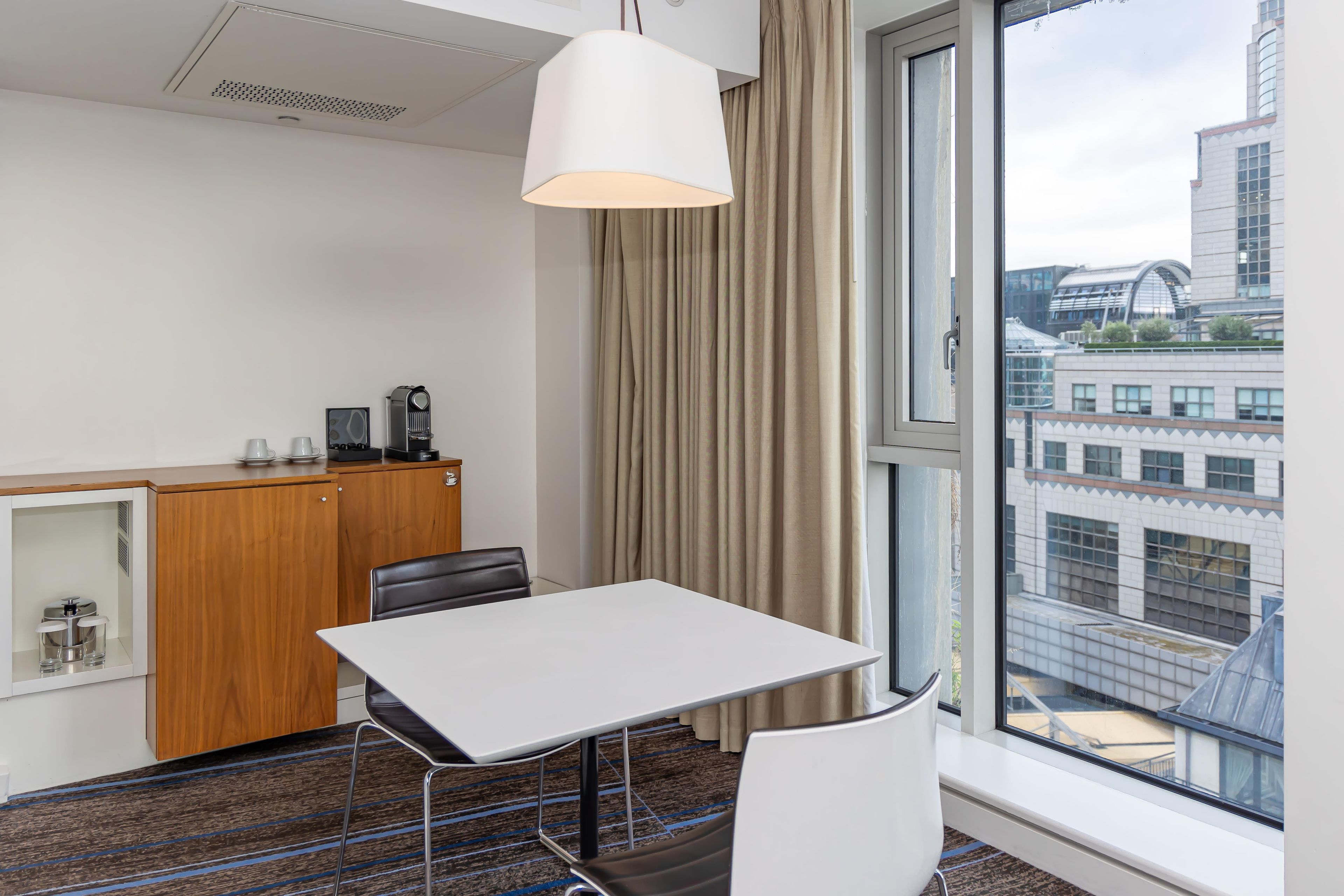 Une chambre d’hôtel avec une grande fenêtre, une table, des chaises, un bureau avec une machine à café et un rideau.