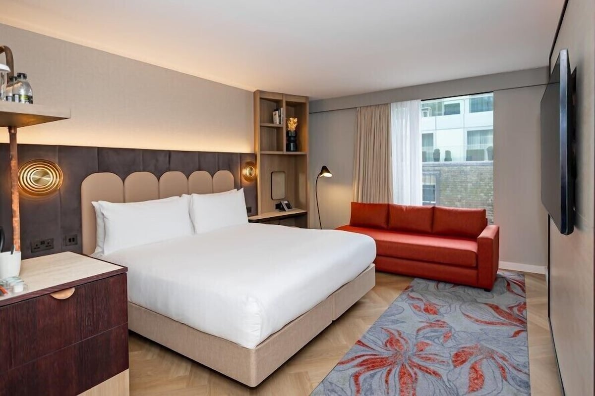 A hotel room with a large bed, a red sofa, a TV, a desk with a lamp, and a window with curtains.