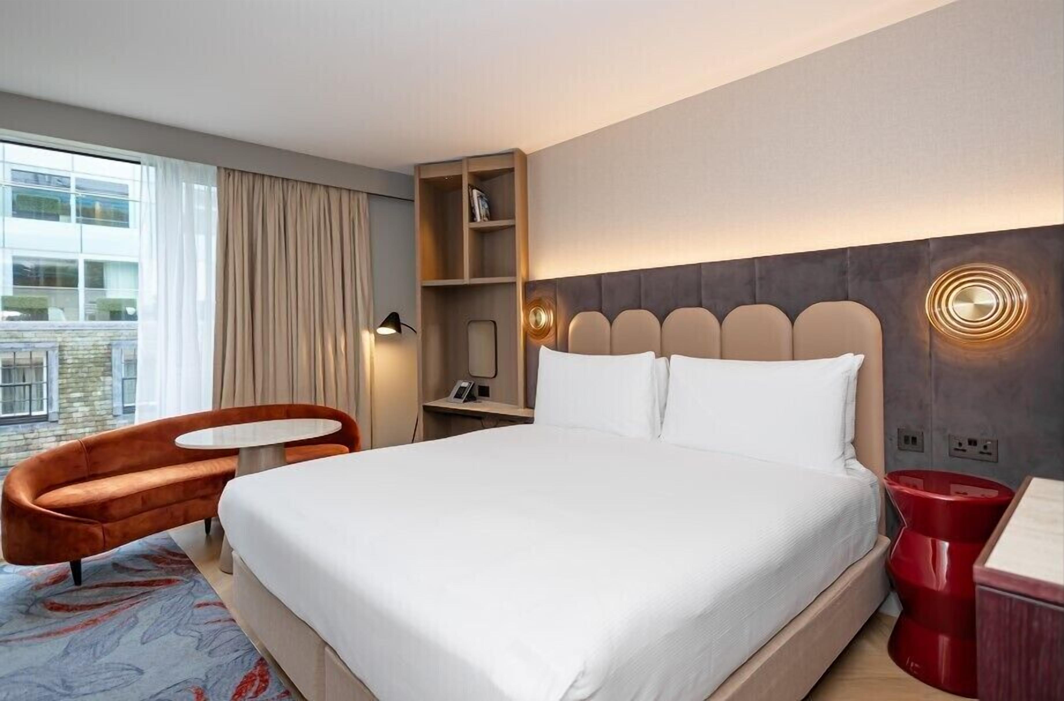 A hotel room with a large bed, a small round table, a sofa, a bookshelf, and a window with curtains.