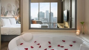 A hotel room with a large window, a bathtub with rose petals, a bed, and a city view. - Sivatel Bangkok (Bangkok)