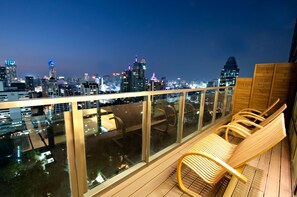 A balcony with wooden chairs and a glass railing overlooking a cityscape at night. - Sivatel Bangkok (Bangkok)