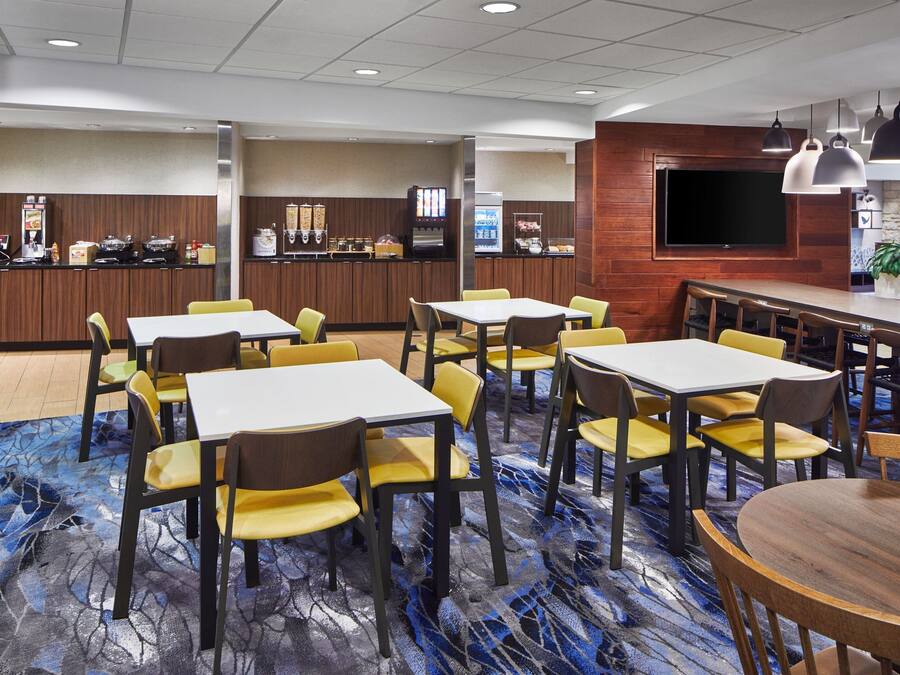 A hotel dining area with tables and chairs, a counter with a beverage dispenser, and a television mounted on the wall.